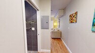 Located at 360 Main St in Hartford, CT, this efficiently designed single-floor unit makes excellent use of its compact footprint. The open-plan main area seamlessly integrates a living space and a fully equipped kitchen, creating a functional and cohesive environment. The kitchen features contemporary light wood cabinetry, dark countertops, stainless steel appliances including a refrigerator and microwave, and a sleek glass backsplash. Track lighting runs along the ceiling, providing ample illumination throughout. A wall-mounted flat-screen TV and a fold-down dining table maximize the use of available space. Warm hardwood flooring extends throughout the main living area, complementing the neutral, light grey walls. Built-in overhead storage cabinets and modular shelving units offer smart organizational solutions, while colorful abstract artwork adds personality to the space. The bathroom is a standout feature, finished floor-to-ceiling in large-format dark grey tiles accented by a decorative mosaic tile border. It includes an open, roll-in style shower area with a curved curtain rod, a wall-mounted grab bar, and stone-look floor tiles, suggesting accessibility-conscious design. A hallway connects the living area to the bathroom, with an electrical panel conveniently recessed into the wall. Overall, the unit presents a clean, modern aesthetic with practical, space-conscious design throughout.