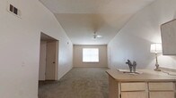 This single-floor residential space offers a clean, functional layout designed with comfort in mind. The main living area is notably spacious, featuring soft carpeted flooring that extends throughout the room, contributing to a warm and inviting atmosphere. Vaulted ceilings rise above, adding a sense of openness and volume to the interior. A ceiling fan is centrally mounted, providing both practicality and a subtle design element overhead. Natural light flows generously into the space through a centrally positioned window, brightening the neutral-toned walls and giving the room an airy, welcoming feel. The walls are painted in a soft, neutral beige, creating a versatile backdrop well-suited to a variety of furnishing styles. A wooden dresser or cabinet is visible along one side, suggesting a bedroom or multi-use living space. Doorways on either side of the main room lead to additional areas, hinting at a connected layout with accessible adjoining rooms or closets. The overall ambiance is simple, bright, and move-in ready, offering a blank canvas for personalization.