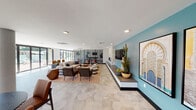 This expansive single-floor space combines modern design with rich cultural influences to create a dynamic and inviting communal environment. The open layout is anchored by large-format hexagonal floor tiles in a warm neutral tone, lending a cohesive and sophisticated foundation throughout.

The main lounge area features multiple flexible seating arrangements, including plush sofas, leather accent chairs, and patterned ottomans arranged atop textured area rugs. A striking black laser-cut decorative screen partition with an intricate geometric Moorish motif divides the lounge from an adjacent workspace or game area, adding both visual interest and functional zoning without fully closing off the space.

One accent wall is painted a soft teal and showcases a large-scale framed artwork depicting an ornate Moorish architectural arch, complemented by a built-in media console and a sleek linear fireplace set into a stone-clad wall. A bistro-style dining nook with high-top tables and bar stools lines another wall, adorned with a gallery of framed patterned prints beneath pendant lighting.

The kitchen area is finished with a quartz waterfall island, arabesque tile backsplash, wood cabinetry, and pendant lighting. Floor-to-ceiling windows and glass doors invite abundant natural light and provide views of an outdoor area, enhancing the overall airy and open ambiance.