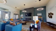 This contemporary single-floor communal space makes a striking first impression with its soaring ceilings and herringbone-patterned hardwood flooring that flows throughout. The welcoming entryway, adorned with curated sculptural wall art, leads into a spacious open-concept layout designed for both relaxation and social interaction.

The lounge area is anchored by a floor-to-ceiling stacked-stone fireplace fitted with a sleek linear hearth and a wall-mounted flat-screen television above. Vaulted ceilings accentuate the vertical drama of the space, while bold teal accent walls and geometric wood paneling add depth and character. Circular halo pendant lights hang overhead, casting a warm, modern glow across the seating arrangement of vibrant blue channel-tufted sofas and coordinating gray velvet upholstered pieces, all accented with amber and rust throw pillows.

An adjacent dining and kitchen area provides a functional yet equally stylish counterpart to the lounge. A large dark-topped island with bold pendant dome lights in black and gold serves as the focal point, complemented by rich teal cabinetry, a herringbone-tile backsplash, and bar seating with orange accent chairs. Multiple round dining tables with leather-style chairs are arranged nearby, all bathed in natural light from large glass-panel doors that open to views of the surrounding exterior.