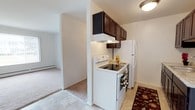 This single-floor residence offers a practical and well-organized layout designed for comfortable everyday living. The kitchen serves as a central functional hub, featuring dark espresso-toned upper cabinetry that provides ample storage space. White appliances, including an electric range with an overhead exhaust hood and a full-size refrigerator, are neatly arranged along the kitchen wall. The flooring in the kitchen consists of light-toned tiles, adding a clean and easy-to-maintain surface underfoot, complemented by a decorative area rug near the appliances. A structural column defines the boundary between the kitchen and the adjacent living area, creating a natural transition between the two spaces. The living area is finished with plush wall-to-wall carpeting in a neutral tone, lending warmth and comfort to the room. A baseboard heater along the far wall of the living area ensures efficient climate control throughout the colder months. The walls throughout are finished in a clean, neutral white, reflecting light and contributing to an open, airy ambiance. Flush-mount ceiling lighting provides consistent illumination across the space. Overall, the unit presents a straightforward, functional design well-suited for daily living.