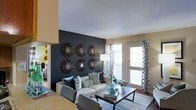 This single-story living space makes a strong visual impression through its thoughtfully designed interior. The living room serves as the focal point, anchored by a dramatic dark accent wall adorned with a symmetrical arrangement of six ornate sunburst mirrors, adding depth and artistic character to the room. Neutral-toned walls throughout the space complement this bold design choice, creating a balanced contrast.

The living area is comfortably sized, featuring hardwood-style flooring that adds warmth underfoot. A light-colored sectional sofa with an array of patterned throw pillows provides ample seating, while a rectangular wood-and-metal coffee table grounds the space. A light blue area rug ties the seating arrangement together cohesively.

Natural light filters into the room through a large window and an exterior door dressed with patterned drapes, offering both brightness and privacy. The door provides direct outdoor access, enhancing the functionality of the space. A glimpse into an adjacent room reveals additional living areas, suggesting a connected, open layout throughout the single-floor home. Overall, the space balances bold design elements with comfortable, practical living.