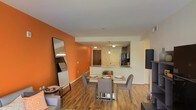 This single-floor space makes a striking first impression with its bold orange accent wall, which anchors the open-concept living and dining area with a warm, energetic ambiance. Rich hardwood-style flooring runs throughout, lending continuity and warmth to the entire space.

The dining area is centrally positioned and features a sleek glass-top table with contemporary gray upholstered chairs, making it an inviting spot for everyday meals or entertaining. A large floor-length mirror with a black frame leans against the orange accent wall, adding depth and dimension to the room.

The kitchen is seamlessly integrated into the layout via a pass-through counter with a light stone surface, creating an open connection between the cooking and dining spaces. Pendant lighting hangs above the counter, providing focused illumination with a modern aesthetic. The kitchen itself features dark cabinetry, a tile backsplash, and standard appliances, offering a functional and stylish cooking environment.

Natural light filters through the space, complemented by recessed and pendant lighting fixtures that keep the interior bright and welcoming. The overall design balances bold color choices with neutral tones and modern furnishings, resulting in a cohesive, contemporary living environment that feels both practical and visually dynamic.