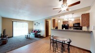 This single-floor space is designed around an open-concept floor plan that seamlessly connects the living, dining, and kitchen areas into one cohesive living environment. The transition between spaces is defined by a change in flooring, with hardwood-style planks in the dining and kitchen areas giving way to carpet in the living room, adding warmth and definition to each zone.

The living area is well-lit by a sliding glass door with vertical blinds, allowing natural light to filter in. A ceiling fan with an integrated light fixture serves as a practical focal point overhead. The kitchen features medium-toned wooden cabinetry, a granite-style countertop, and a built-in wine rack above the upper cabinets. A raised breakfast bar separates the kitchen from the dining area and is illuminated by a multi-light pendant fixture. Black appliances complete the kitchen's functional design.

A bistro-style high-top table with bar chairs occupies the dining space, making efficient use of the area between the kitchen and living room. A doorway visible in the background leads to additional rooms. The textured ceiling and neutral wall tones contribute to the space's understated, functional character.