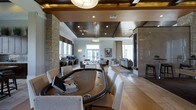 The resident lounge is a thoughtfully designed, single-floor amenity space that blends entertainment and relaxation into one cohesive environment. The interior showcases a mix of materials and textures, including stacked stone accent columns, rich dark wood ceiling panels with recessed lighting, and light tile flooring that unifies the space. A prominent poker table with padded seating anchors the foreground, making it a centerpiece for social gatherings and game nights. Moving further into the space, a foosball table provides additional recreational options, while a designated computer workstation area along one wall offers practical connectivity for residents. The lounge area transitions seamlessly into a comfortable seating zone furnished with contemporary sofas and chairs, ideal for casual conversation or unwinding. Large windows and glass doors allow natural light to filter throughout, enhancing the open and inviting atmosphere. Pendant lighting and wall sconces complement the recessed fixtures, creating a warm ambiance during evening hours. The overall design strikes a balance between sophisticated aesthetics and functional amenities, offering residents a versatile space suited for both lively entertainment and quiet relaxation.
