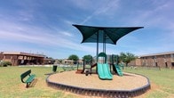 This outdoor playground area offers a thoughtfully designed recreational space suitable for children of various ages. At its centerpiece stands a multi-level play structure finished in teal green, featuring two slides of varying sizes — including a tunnel slide and an open slide — alongside climbing ladders, handrails, and interactive panel elements such as spinners and tactile activity boards. The entire structure sits within a defined, edged border filled with wood chip surfacing, providing a cushioned, safety-conscious ground cover. Overhead, a large dark green shade canopy with a distinctive spread-wing design provides generous protection from direct sunlight, making the space comfortable even on bright, sunny days. Surrounding the playground is a well-kept grassy lawn, with additional seating available via nearby benches. A swimming pool is visible in the background, suggesting this playground is part of a broader recreational amenity area. The space is enclosed by low fencing and flanked by brick residential buildings, creating a contained and secure environment. The open sky above and the lush greenery around the perimeter lend the area a bright, welcoming ambiance ideal for active outdoor play.