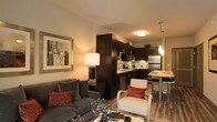 This single-floor residence showcases an open-concept floor plan that seamlessly integrates the living, dining, and kitchen areas into one cohesive space. The living room is anchored by a plush gray sofa accented with red throw pillows, a sleek glass coffee table, and a framed abstract artwork that adds a sophisticated touch to the warm-toned walls. A contemporary floor lamp provides soft ambient lighting, enhancing the inviting atmosphere.

The kitchen is outfitted with dark espresso cabinetry, stainless steel appliances, and a mosaic tile backsplash that adds visual texture. The workspace is practical and well-organized, with ample counter space and under-cabinet storage. A pendant light hangs above a raised bar-height dining table, creating a natural transition between the kitchen and the rest of the living space. Bar-style seating further reinforces the open, social layout.

Hardwood-style flooring extends throughout, contributing warmth and continuity across the space. The overall design palette — combining dark cabinetry, neutral walls, and bold accent colors — creates a modern, urban aesthetic that is both functional and visually appealing.