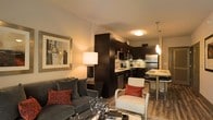 This single-floor residence showcases an open-concept floor plan that seamlessly integrates the living, dining, and kitchen areas into one cohesive space. The living room is anchored by a plush gray sofa accented with red throw pillows, a sleek glass coffee table, and a framed abstract artwork that adds a sophisticated touch to the warm-toned walls. A contemporary floor lamp provides soft ambient lighting, enhancing the inviting atmosphere.

The kitchen is outfitted with dark espresso cabinetry, stainless steel appliances, and a mosaic tile backsplash that adds visual texture. The workspace is practical and well-organized, with ample counter space and under-cabinet storage. A pendant light hangs above a raised bar-height dining table, creating a natural transition between the kitchen and the rest of the living space. Bar-style seating further reinforces the open, social layout.

Hardwood-style flooring extends throughout, contributing warmth and continuity across the space. The overall design palette — combining dark cabinetry, neutral walls, and bold accent colors — creates a modern, urban aesthetic that is both functional and visually appealing.