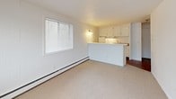 This single-floor, 1-bedroom, 1-bathroom unit offers a straightforward and functional layout designed for comfortable everyday living. The open-concept living and dining area features neutral carpeting, white panel walls, and a paneled ceiling, creating a clean and bright atmosphere. A window along the exterior wall allows natural light to filter in, while baseboard heating and a wall-mounted air conditioning unit ensure year-round climate control.

The kitchen is accessible directly from the living area via a half-wall peninsula counter, offering an open pass-through feel. It is equipped with white cabinetry — both upper and lower — a gas range with overhead ventilation, a refrigerator, and a stainless steel single-basin sink set into stone-patterned laminate countertops. The hardwood-style flooring in the kitchen adds a warm contrast to the carpeted living spaces.

The bedroom is a generously sized, carpeted room with white walls, a paneled ceiling, and a window providing natural light and views of the surrounding area. Baseboard heating runs along the exterior wall.

The bathroom features full white subway tile walls, a classic mosaic tile floor with decorative accents, a built-in tub and shower, a wood-toned vanity with an integrated sink, and a mirrored medicine cabinet above for added storage.