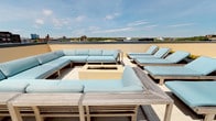 This rooftop terrace offers an elevated outdoor retreat with sweeping views of the surrounding skyline and tree canopy. The centerpiece of the space is a built-in rectangular fire pit, finished in a warm neutral tone, and framed by generously sized sectional sofas outfitted with light blue weather-resistant cushions and a natural wood frame. The seating arrangement creates an intimate, lounge-like atmosphere ideal for social gatherings. To one side, a row of matching teak-framed chaise lounges with coordinating cushions provides a dedicated sunbathing area. The open-air layout allows for abundant natural light throughout the day, while the unobstructed sightlines to the horizon enhance the sense of openness and space. The clean, modern aesthetic is carried through the consistent use of neutral tones, warm wood finishes, and cohesive furnishings. Whether used for relaxing in the sun or gathering around the fire at dusk, this rooftop terrace functions as a versatile outdoor amenity with a comfortable and well-appointed design.