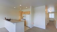 This two-floor space offers a clean, functional layout designed for everyday comfort. The living area is carpeted and bathed in natural light from multiple sliding windows that frame views of the surrounding neighborhood. Baseboard heating runs along the exterior walls, providing efficient climate control throughout the room. Neutral cream-toned walls and white ceilings contribute to a bright, airy atmosphere, while recessed lighting in select areas supplements natural light.

The kitchen is open to the main living space and features light wood cabinetry with ample upper and lower storage. White appliances, including a gas range, over-the-range microwave, and a full-size refrigerator, are included. Recessed lighting illuminates the kitchen area effectively. A partial countertop bar with a raised edge separates the kitchen from the living space, creating a natural transition between the two areas.

The carpeted floors extend throughout the main living zone, giving the space a cohesive, unified feel. The overall design is simple and well-maintained, emphasizing practicality and light, with a neutral palette that offers flexibility for personalization across both floors.
