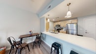 Located at 696 Whitney Rd W in Fairport, NY, this third-floor apartment offers a well-designed, single-level layout that maximizes both comfort and functionality. The open-concept living and dining area is bathed in soft, neutral tones with light sage-green walls and durable wood-look flooring that flows seamlessly throughout the main living spaces. A sliding glass door in the living area provides access to an outdoor balcony, allowing natural light to fill the room. The kitchen is efficiently designed with light maple cabinetry, stainless steel appliances including a refrigerator, electric range, and microwave, and a white countertop peninsula that doubles as a breakfast bar with pendant lighting overhead — ideal for casual dining or entertaining. The bedroom features plush carpeting, multiple windows with natural light, and ample space for a full furniture arrangement. The bathroom is finished with a wood-toned vanity, a large mirror with overhead lighting, and a walk-in shower with grab bar accessibility. Throughout the unit, clean lines, neutral palettes, and practical finishes create a calm, cohesive atmosphere well-suited for comfortable everyday living in the Fairport community.