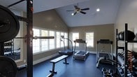 This single-floor fitness space is thoughtfully designed to support a comprehensive workout routine. The room's most striking architectural feature is its vaulted ceiling, which rises to a pronounced peak and lends an open, airy quality to the space. A ceiling fan is centrally mounted, providing ventilation and air circulation during workouts.

Natural light floods the room through multiple windows positioned along the walls, offering views of the surrounding greenery and creating a bright, motivating atmosphere. The warm taupe-toned walls complement the dark rubber flooring, which is well-suited for heavy equipment use and provides both durability and traction underfoot.

The space is outfitted with a Rogue power rack mounted along one wall, allowing for strength training exercises including barbell work. A flat weight bench and a set of dumbbells accompany the rack for versatile free weight training. Two treadmills are positioned toward the center and far end of the room, offering dedicated cardio options. Additional fitness equipment rounds out the setup near the far wall. Recessed lighting supplements the natural light, ensuring the space remains well-lit during any time of day.