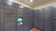 This dedicated package room at Waterside Village is designed with resident convenience at its core. The space is outfitted with an extensive Amazon Hub smart locker system that spans multiple walls in an L-shaped configuration, providing a large number of individual compartments to accommodate deliveries of various sizes. The lockers feature a centrally mounted interactive touchscreen kiosk with a keypad, enabling residents to retrieve packages securely at any time of day.

The room itself is finished to a clean, functional standard, with neutral cream-toned walls and rich dark hardwood-style flooring that lends a polished look to an otherwise utilitarian space. Recessed lighting overhead ensures the room is well-lit and easy to navigate. A patterned bench near the entrance offers a convenient spot to manage packages upon retrieval, and a branded Waterside Village entry mat anchors the doorway. Subtle decorative touches, including a large abstract canvas artwork mounted on the wall, elevate the ambiance beyond a typical utility room. The layout is thoughtfully organized, with clear signage indicating locker assignments for all units. Overall, the package room reflects a modern, resident-focused amenity that prioritizes both security and ease of use.