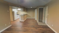 This single-floor space offers an open-concept layout that seamlessly connects the kitchen and living area, creating a practical and versatile floor plan. The kitchen is positioned along one wall and features white cabinetry, a white refrigerator, a white range, and dark countertops. A peninsula with a raised bar-height counter extends outward, providing additional workspace and a natural division between the kitchen and the open living area. The flooring throughout the main living space consists of rich, dark hardwood-style planks that add warmth and visual continuity. Neutral tan walls provide a clean, understated backdrop that complements the flooring. A wall-mounted HVAC unit is visible near the kitchen area, offering climate control. Recessed lighting and under-cabinet fluorescent lighting illuminate the kitchen zone effectively. Toward the back of the space, an open walk-in closet offers generous storage, while an adjacent doorway leads to a tiled area, likely a bathroom or utility space. A textured ceiling adds subtle architectural interest. Overall, the space presents a straightforward, functional layout with clean finishes suitable for comfortable everyday living.