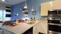 This single-floor space showcases a thoughtfully designed open-concept layout where the kitchen and living area flow seamlessly together. The kitchen is a standout feature, equipped with crisp white cabinetry, light granite countertops, and a subway tile backsplash in a soft blue-gray tone that complements the bold blue accent walls throughout. A stainless steel sink is centrally positioned along the kitchen peninsula, which doubles as a functional breakfast bar and social hub. Overhead, sleek teardrop-style pendant lights hang at varying heights, adding a modern, artistic touch to the space. The open shelving mounted on the blue wall provides both display and storage opportunities, while contributing to the airy, contemporary feel of the interior. The living area, visible from the kitchen, features warm, layered décor with cushioned seating and accent lighting, creating an inviting atmosphere. The consistent use of blue tones across the walls ties the kitchen and living spaces together cohesively. The overall ambiance is modern and vibrant, balancing bold color choices with clean, minimalist design elements for a stylish and functional living environment.