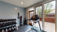 This single-level fitness center is thoughtfully designed to support a full range of workout routines. The space is finished with durable rubber flooring throughout, providing both safety and comfort during exercise. Soft blue-gray walls and recessed lighting create a clean, focused atmosphere conducive to training.

The cardio zone is well-appointed, featuring a treadmill, elliptical trainer, and upright stationary bike, all positioned near large sliding glass doors and windows that flood the area with natural light and offer views of an adjacent outdoor patio. A wall-mounted flat-screen TV is positioned for convenient viewing during cardio sessions.

The strength training area includes a multi-functional cable machine system, a fully stocked dumbbell rack with a wide range of weights, a medicine ball rack with resistance bands, kettlebells, a stability ball, and an adjustable workout bench. These elements together accommodate both free weight and machine-based training.

A full-length mirror is mounted near the entry, supporting form checks during exercise. The space also provides access to a restroom and connects via a corridor to additional amenities. A ceiling-mounted HVAC unit ensures a comfortable environment year-round. Direct access to the outdoor patio further enhances the overall fitness experience.