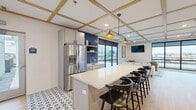 This well-appointed single-floor amenity space is designed for both social gatherings and relaxed leisure. A defining architectural feature is the wood-trimmed coffered ceiling that runs throughout, lending a refined, contemporary character to the entire space. Warm-toned pendant lights hang above the central kitchen island, which is topped with white quartz and lined with dark wood bar stools, providing ample seating for socializing.

The kitchenette area is anchored by a bold navy glazed brick tile backsplash, complemented by gray cabinetry, open wood shelving, a stainless steel refrigerator, and a single-basin sink. Geometric black-and-white patterned floor tile contrasts with the light wood-look flooring found throughout the remainder of the space.

The lounge area is bright and open, benefiting from floor-to-ceiling windows on multiple walls that offer expansive outdoor views and flood the interior with natural light. Comfortable seating, a mounted flat-screen television, and a checkered area rug furnish this zone. Adjacent to the lounge, an open game area accommodates a foosball table, completing the space's recreational offerings.
