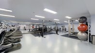 This fitness facility offers a spacious, purpose-built environment designed to support a full range of strength and conditioning activities. The space is finished with clean white flooring that transitions to rubberized, dark-toned flooring in dedicated equipment zones, providing both durability and safety during workouts. Recessed rectangular panel lighting runs along the ceiling, delivering consistent, even illumination throughout the entire floor. The generous ceiling height adds to the open and airy atmosphere of the room. A full-length mirror spans one wall, enhancing the sense of depth and allowing users to monitor their form during exercise. The strength training area is comprehensively outfitted with cable machines, squat racks, weight benches, free weights, and a variety of resistance machines to accommodate diverse training needs. Informational charts and posters are mounted along the walls, providing guidance and reference for users. The open layout allows for ample circulation space between equipment stations, ensuring a comfortable and functional workout environment. The overall design is clean, professional, and practical, reflecting a facility built with performance and usability in mind.