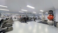 This fitness facility offers a spacious, purpose-built environment designed to support a full range of strength and conditioning activities. The space is finished with clean white flooring that transitions to rubberized, dark-toned flooring in dedicated equipment zones, providing both durability and safety during workouts. Recessed rectangular panel lighting runs along the ceiling, delivering consistent, even illumination throughout the entire floor. The generous ceiling height adds to the open and airy atmosphere of the room. A full-length mirror spans one wall, enhancing the sense of depth and allowing users to monitor their form during exercise. The strength training area is comprehensively outfitted with cable machines, squat racks, weight benches, free weights, and a variety of resistance machines to accommodate diverse training needs. Informational charts and posters are mounted along the walls, providing guidance and reference for users. The open layout allows for ample circulation space between equipment stations, ensuring a comfortable and functional workout environment. The overall design is clean, professional, and practical, reflecting a facility built with performance and usability in mind.