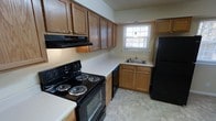 This single-floor residence offers a practical and well-appointed layout designed for comfortable everyday living. The kitchen serves as a central feature of the space, showcasing warm honey-toned oak cabinetry that extends both above and below the countertops, providing generous storage throughout. The kitchen is equipped with a full suite of black appliances, including a coil-burner electric range with oven, a built-in dishwasher, and a full-sized refrigerator. A under-cabinet range hood is positioned above the stove for efficient ventilation. The countertops are finished in a clean, light-colored surface that contrasts nicely with the rich wood tones of the cabinetry. The sink is situated beneath a window that allows natural light to brighten the space, creating an inviting atmosphere. The kitchen floor is laid with neutral tile in a classic pattern, adding a subtle texture underfoot. The layout follows an efficient galley-style configuration, making excellent use of the available space by positioning appliances and workspaces along two parallel walls. Overall, the space presents a tidy, functional character with a warm, traditional aesthetic that makes it well-suited for daily use.