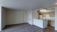 This single-floor unit presents an open-concept design that connects the living area to the kitchen, creating a cohesive and functional layout. The living area is finished with wall-to-wall carpet in a neutral gray tone, providing a comfortable underfoot feel, while the kitchen and adjacent spaces transition to hard flooring, offering a practical contrast.

The kitchen is positioned at one end of the open layout and features a raised breakfast bar that serves as a natural divider between the living and cooking areas. Wood-toned upper cabinetry lines the kitchen walls, complemented by a white refrigerator and light countertops. An overhead fluorescent light fixture provides direct illumination to the kitchen workspace.

The walls throughout are painted in a clean, neutral off-white, contributing to a bright and airy atmosphere. HVAC vents are integrated into both the walls and ceiling, ensuring climate control throughout the space. A standard interior door provides access to additional areas of the unit. The overall ambiance is clean, minimal, and move-in ready, suited to a variety of furnishing styles.