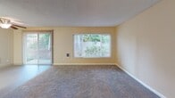 This single-floor residence offers a well-organized layout designed for comfortable everyday living. The expansive living room is a standout feature, finished with plush carpet and warm, light yellow walls that create a welcoming ambiance. Large windows fitted with horizontal blinds flood the space with natural light and offer views of the surrounding greenery, while a sliding glass door with vertical blinds provides direct access to an outdoor patio area.

The kitchen is laid out in a practical galley style, featuring warm-toned wood cabinetry with ample upper and lower storage, white countertops, a white electric range, a sink, and a full-size refrigerator. Light vinyl flooring completes the clean, functional look of this space.

The bedrooms are carpeted and finished with the same warm neutral tones found throughout the home. The primary bedroom includes a walk-in closet and a window that brings in natural light. An additional bedroom offers generous proportions with a ceiling light fixture.

The bathroom is neatly appointed with white square-tiled walls surrounding a full soaking tub and shower combination, chrome fixtures, and light wood-look flooring. Textured ceilings are present throughout the home, adding a consistent architectural detail, while white baseboards provide clean contrast against the neutral wall palette.