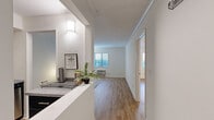 This one-bedroom, one-bathroom unit at River Hill Flats offers a efficiently designed single-floor layout finished with light-toned wood flooring throughout. The open-concept kitchen and living area form the heart of the unit, with a hallway connecting the spaces seamlessly. The kitchen is outfitted with dark cabinetry, stainless steel appliances including a refrigerator, electric range, built-in microwave, and a stainless steel sink with ample counter space. A raised counter divides the kitchen from the main corridor, providing functional separation without closing off the space. The living room receives natural light through a window with exterior views, and features a wall-mounted HVAC unit. Crown molding adds a clean finishing detail along the ceiling line. The bedroom is a comfortable size, complete with a ceiling fan, light wood flooring, and direct sightlines to the kitchen area. The bathroom features a dark vanity with white countertop, a well-lit mirror, and a shower-tub combination. White walls and consistent flooring throughout give the unit a cohesive, clean aesthetic.