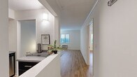 This one-bedroom, one-bathroom unit at River Hill Flats offers a efficiently designed single-floor layout finished with light-toned wood flooring throughout. The open-concept kitchen and living area form the heart of the unit, with a hallway connecting the spaces seamlessly. The kitchen is outfitted with dark cabinetry, stainless steel appliances including a refrigerator, electric range, built-in microwave, and a stainless steel sink with ample counter space. A raised counter divides the kitchen from the main corridor, providing functional separation without closing off the space. The living room receives natural light through a window with exterior views, and features a wall-mounted HVAC unit. Crown molding adds a clean finishing detail along the ceiling line. The bedroom is a comfortable size, complete with a ceiling fan, light wood flooring, and direct sightlines to the kitchen area. The bathroom features a dark vanity with white countertop, a well-lit mirror, and a shower-tub combination. White walls and consistent flooring throughout give the unit a cohesive, clean aesthetic.