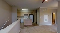 This single-floor residence showcases an open-concept layout that seamlessly connects the kitchen and living areas. The kitchen is equipped with light wood cabinetry offering generous storage, a white refrigerator, a dishwasher, and a range. A central kitchen island provides additional workspace and serves as a natural dividing point between the kitchen and the adjacent living area. The flooring in the kitchen features neutral-toned tile, which transitions into wall-to-wall carpeting in the living space, creating a clear distinction between the two zones. The walls are finished in a warm beige tone throughout, contributing to a cohesive and understated aesthetic. Crown molding is visible along the upper edges of the walls, adding a subtle architectural detail. A ceiling fan is partially visible, suggesting climate comfort features are integrated into the space. A door near the rear of the kitchen leads to an additional room or utility area. Overhead fluorescent lighting illuminates the kitchen, while HVAC vents are positioned along the walls to ensure consistent airflow throughout the unit.