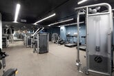 This well-equipped fitness center at 401 Williams Ave SW in Huntsville, Alabama, offers a thoughtfully designed single-floor layout accommodating a wide range of workout needs. The space is divided into distinct functional zones, each tailored to a specific type of training. The main strength training area is expansive and bright, featuring rows of Life Fitness selectorized machines, cable systems, and free weight equipment arranged to allow easy navigation and use. Linear LED lighting runs along the ceiling, providing consistent, even illumination throughout this section and lending the space a clean, modern aesthetic. The flooring throughout is durable rubber gym matting, providing comfort and safety underfoot. A dedicated cardio zone, distinguished by deep blue accent walls and a dark ceiling with recessed lighting, creates a more intimate atmosphere. This area houses treadmills, ellipticals, and other cardiovascular machines, along with a wall-mounted television for entertainment during workouts. A barre rail is also visible along one wall, suggesting additional fitness programming options. Accessible restroom facilities are conveniently located within the facility. The overall design is functional and contemporary, with a neutral and industrial palette in the main area transitioning to a more energized, bold aesthetic in the cardio zone.