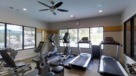 Located at 8000 Waters Ave in Savannah, Georgia, this single-floor fitness center is designed to provide a functional and comfortable workout environment. The space is equipped with a variety of cardio machines, including treadmills, elliptical trainers, and a stationary bike, thoughtfully arranged to allow for easy movement between equipment. Large windows line the walls, flooding the room with natural light and offering pleasant views of the surrounding outdoor greenery, creating an open and inviting atmosphere. A ceiling fan mounted overhead works in tandem with recessed lighting to keep the space well-ventilated and evenly illuminated throughout the day. The neutral color palette, accented by a warm horizontal band along the walls, contributes to a clean and focused aesthetic. Durable flooring with area mats beneath each piece of equipment adds both safety and visual structure to the layout. A wall clock ensures exercisers can easily manage their workout time. The combination of natural light, greenery views, and well-maintained equipment makes this fitness center a practical and appealing space for residents or members looking to maintain an active lifestyle.