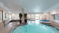 This indoor pool facility offers a refined aquatic environment designed for year-round use. The centerpiece is a generously sized swimming pool with vibrant turquoise water, complemented by a freeform hot tub/spa finished in classic tile work. A central structural column, flanked by tall decorative potted plants, adds an architectural element to the space while visually dividing the pool and spa areas.

The facility is flooded with natural light through large windows and glass-panel doors that line the perimeter, offering clear views of an adjacent outdoor pool area. These doors provide direct exterior access, seamlessly connecting indoor and outdoor aquatic amenities. The tile flooring throughout is both practical and polished, with stepped platforms providing safe and accessible transitions between different levels of the space.

The interior walls are finished in clean white, accented by framed artwork that adds a touch of sophistication to the utilitarian setting. Recessed and flush-mounted ceiling lights supplement the natural light, ensuring the space is well-lit at all times. Safety features are prominently displayed, including posted pool rules and fire safety equipment. An elevator and multiple doorways provide convenient access throughout. The overall ambiance is bright, clean, and inviting, reflecting a well-maintained recreational facility.