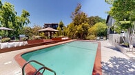 Located at 949 East St in Lafayette, CA, this outdoor amenity space offers a well-appointed communal retreat within a multi-building residential community. At the heart of the space is a generously sized swimming pool with clear turquoise water, bordered by a classic brick-tiled coping and a spacious concrete pool deck that provides ample room for lounging and recreation. Adjacent to the pool is a spa, also finished with matching brick tile detailing, completing the resort-style atmosphere. The pool area is enclosed by sleek black wrought iron fencing, ensuring both security and an open, airy feel. Surrounding the space, mature deciduous trees provide natural shade and a sense of privacy, while carefully maintained landscaping with ornamental plants and garden beds adds color and texture to the environment. A raised lounge terrace equipped with seating and umbrellas offers an additional gathering area perfect for relaxing outdoors. The multi-story residential buildings framing the courtyard feature contemporary gray lap siding, wood-accented balconies, and large windows that overlook the pool area. Concrete stairways with black metal railings provide accessible connections between levels of the complex. Under the abundant California sunshine, this outdoor space captures a serene, community-focused ambiance ideal for leisure and social enjoyment.
