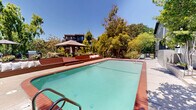 Located at 949 East St in Lafayette, CA, this outdoor amenity space offers a well-appointed communal retreat within a multi-building residential community. At the heart of the space is a generously sized swimming pool with clear turquoise water, bordered by a classic brick-tiled coping and a spacious concrete pool deck that provides ample room for lounging and recreation. Adjacent to the pool is a spa, also finished with matching brick tile detailing, completing the resort-style atmosphere. The pool area is enclosed by sleek black wrought iron fencing, ensuring both security and an open, airy feel. Surrounding the space, mature deciduous trees provide natural shade and a sense of privacy, while carefully maintained landscaping with ornamental plants and garden beds adds color and texture to the environment. A raised lounge terrace equipped with seating and umbrellas offers an additional gathering area perfect for relaxing outdoors. The multi-story residential buildings framing the courtyard feature contemporary gray lap siding, wood-accented balconies, and large windows that overlook the pool area. Concrete stairways with black metal railings provide accessible connections between levels of the complex. Under the abundant California sunshine, this outdoor space captures a serene, community-focused ambiance ideal for leisure and social enjoyment.