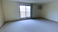 This single-floor space offers a straightforward and functional layout designed for comfortable everyday living. The main living area is finished with wall-to-wall carpeting in a neutral tone, providing a soft underfoot feel and a clean, understated aesthetic throughout. A large sliding glass door serves as the room's focal point, allowing natural light to fill the interior while providing direct access to an outdoor balcony enclosed by a classic white railing. The expansive glazing frames exterior views and contributes to an open, airy atmosphere within the room. Climate comfort is addressed through a combination of a wall-mounted air conditioning unit positioned near the upper wall and baseboard heating that runs along the perimeter of the room, ensuring year-round temperature control. The walls are finished in a light, neutral color that enhances the brightness of the space. Vertical window treatments offer adjustable privacy and light filtration. The overall design is clean and minimal, making the space highly adaptable for a variety of furnishing arrangements and personal styles. This unit presents a practical and livable environment suited for those seeking a no-frills, move-in ready space with essential amenities in place.