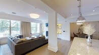 This single-floor residence showcases a thoughtfully designed open-concept layout that connects the living, dining, and kitchen areas in a cohesive and functional arrangement. The living area is anchored by a comfortable sectional sofa and features a large window dressed with floor-length neutral drapes, allowing natural light to filter in while maintaining a soft, warm ambiance. A flush-mount ceiling fixture provides additional illumination, complementing the recessed lighting throughout.

A structural column with clean white millwork detailing serves as a subtle architectural divider between spaces without disrupting the open flow. Built-in shelving units with integrated lighting flank a central media area, offering both display space and a refined decorative focal point. The cabinetry is finished in crisp white, contributing to the overall bright and airy feel of the interior.

Light hardwood flooring runs throughout, unifying the space and adding warmth. A granite countertop edge visible in the foreground hints at a well-appointed kitchen nearby, while a glimpse of a dining area with dark furniture adds contrast. The overall aesthetic is modern and refined, with a neutral palette of whites, grays, and natural wood tones.