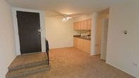 This single-floor unit offers a straightforward, functional layout with an open-concept design that seamlessly connects the living area and kitchen. The living space is finished with wall-to-wall carpeting in a warm neutral tone, providing a comfortable underfoot feel throughout. A ceiling fan with an integrated light fixture is centrally positioned, offering both ventilation and ambient lighting to the main living area.

The kitchen occupies a dedicated alcove along one wall, featuring light wood cabinetry that extends to the upper portion of the wall for generous storage. A dark countertop runs the length of the kitchen, providing practical workspace, and a sink is positioned beneath the upper cabinets. The kitchen flows naturally into the living space without visual obstruction, contributing to the open feel of the unit.

A dark-toned entry door with a brass-finished deadbolt marks the main entrance, situated in the corner of the living area. Adjacent to the entry, a slatted bi-fold door suggests a closet for additional storage. The walls are finished in a clean, neutral off-white, keeping the space feeling bright and versatile. Overall, the unit presents a compact yet efficiently organized single-level living environment suited for everyday functionality.
