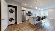 The Maple is a thoughtfully designed two-bedroom, two-bathroom residence located in Ann Arbor, Michigan. The single-floor layout offers a comfortable and functional living experience with contemporary finishes throughout. The open-concept kitchen serves as the heart of the home, featuring white shaker cabinetry, a stainless steel appliance suite, a mosaic tile backsplash, and a large center island with bar seating — all illuminated by sleek pendant lighting. Warm wood-tone flooring flows through the main living areas, adding a welcoming sense of continuity. A dedicated in-unit laundry closet with a stacked washer and dryer provides added convenience. Both bedrooms are carpeted and filled with natural light from well-placed windows. The primary bedroom includes a stylish sliding barn door that opens to a walk-in closet, adding a modern architectural touch. The bathrooms are appointed with dark-finish vanities, white countertops, and tile-surround showers. Neutral gray wall tones throughout the unit create a clean, versatile backdrop that complements the modern design. The Maple combines practical amenities with refined details, making it a well-appointed home in a vibrant Ann Arbor location.