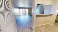 Located at 6303 Indian School Rd NE in Albuquerque, New Mexico, this efficient single-floor unit offers a well-organized layout across a compact footprint. The combined kitchen and living room serves as the heart of the space, featuring light wood-look flooring throughout and a pass-through counter that visually connects the kitchen to the living area. The kitchen is outfitted with gray shaker-style cabinetry extending to the ceiling for ample storage, light countertops, and stainless steel appliances including a range, over-the-range microwave, and full-size refrigerator. The living area benefits from a large sliding glass door dressed with vertical blinds, allowing abundant natural light to fill the room and providing access to an outdoor space with a decorative screen enclosure. A ceiling fan adds comfort overhead. The unit includes two bathrooms: one features a full tub-shower combination with clean white tile surround and chrome fixtures, while the second offers a vanity with stone-look countertops, gray cabinetry, a framed mirror, and a louvered-door closet with wood-look flooring. The entrance and hallway provide a functional transition between spaces, maintaining the consistent neutral color palette and wood-look flooring found throughout the unit.