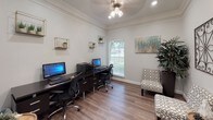 This single-floor space functions as a polished business center designed to support productivity while maintaining a warm, inviting atmosphere. The primary workstation area is anchored by a large dark-wood desk accommodating two desktop computers and a multifunction printer, making it well-suited for shared use. Ergonomic rolling chairs provide comfortable seating, while a window with blinds allows natural light to brighten the room. Decorative gold-framed cube shelves displaying small potted plants and minimalist accents add a refined touch to the neutral white walls. Crown molding and a ceiling fan with integrated lighting further elevate the room's classic character, and wood-look plank flooring runs throughout, lending warmth and cohesion to the space. An adjoining lounge area offers a more relaxed setting, featuring a patterned accent chair, a rustic drum side table, and a striking wrought-iron wall art piece with an ornate scrollwork design. Lush indoor plants punctuate both areas, introducing a natural, refreshing element. Interior glass-paned windows connect the spaces visually, creating an open and airy feel. Together, the rooms strike a balance between functional workspace and comfortable gathering area, making this an ideal environment for residents or visitors needing both work and relaxation amenities.