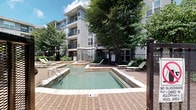 Located at 3949 Lindell Blvd in the heart of St. Louis, MO, this outdoor pool area serves as a central amenity within a multi-story residential community. The space is defined by a clean, rectangular swimming pool with clearly marked depth indicators, accompanied by a shallow wading area — ideal for relaxing in warmer months. The pool deck is finished with classic brick pavers, providing a durable and visually appealing surface that surrounds the water on all sides.

Multiple lounge chairs are positioned around the pool, offering ample space for sunbathing and leisure. Mature trees throughout the courtyard provide natural shade and greenery, contributing to a pleasant outdoor atmosphere. The area is enclosed by a metal security fence with a keyed-access gate, ensuring a private and controlled environment. Signage designates this as a smoke-free and no-glassware zone, reflecting community safety standards.

The pool is set against the backdrop of the multi-story residential building, which features modern gray architecture, private balconies, and large windows. Landscaped garden beds with shrubs and ground cover line the building's exterior, and a concrete walkway connects the pool area to surrounding building entrances, making the space both functional and aesthetically cohesive.