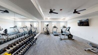 Located at 2155 Arkansas Ln in Grand Prairie, TX, this fitness center offers a well-appointed, single-floor exercise space designed to support a comprehensive workout routine. The room features a tray ceiling with crown molding and is kept comfortable by multiple ceiling fans and flush-mounted lighting fixtures. A full-length wall mirror along one side amplifies the sense of space and allows exercisers to monitor their form.

The cardio section is well-equipped with multiple treadmills, elliptical trainers, a recumbent bike, and a stationary upright bike. A wall-mounted flat-screen television is positioned for convenient viewing during cardio sessions. On the strength training side, the facility includes a multi-function cable machine, an adjustable workout bench, a stability ball, and a full rack of dumbbells spanning a wide range of weights.

The flooring throughout is durable, low-pile carpet in a neutral gray tone, providing traction and cushioning underfoot. Natural light filters in through several windows along the walls, offering views of the surrounding community. The clean white walls and bright, functional lighting create an energizing and practical environment well-suited for daily fitness use.