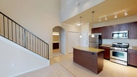 This two-story space immediately impresses with its dramatic, soaring ceilings that amplify the sense of openness throughout the main living area. The kitchen serves as a central focal point, showcasing rich dark wood cabinetry paired with light granite countertops on a curved island that provides both workspace and casual seating. Stainless steel appliances, including a full-size refrigerator, complement the warm cabinetry tones. Pendant lighting hangs above the island, while a track lighting system overhead ensures the kitchen remains well-illuminated. The flooring transitions from light-toned tile in the kitchen and entry areas to plush carpeting in the living space, adding warmth and comfort underfoot. An elegant arched doorway leads to a bathroom or vanity area, visible in the background, contributing to the architectural character of the space. A staircase with a dark metal railing provides access to the upper floor, its clean lines adding a modern touch. The tall walls and open sightlines between levels reinforce the airy, expansive ambiance throughout.