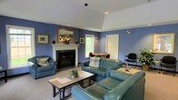 This single-floor space offers a welcoming and well-appointed interior designed for comfort and functionality. The main living area is the centerpiece of the space, characterized by rich blue-painted walls that create a warm, inviting atmosphere. A classic white-mantled fireplace with a dark marble surround serves as the focal point of the room, accented by a gold-framed mirror and floral arrangement on the mantel, along with framed artwork on the surrounding walls. The seating area is furnished with a generously sized teal leather sectional sofa and matching armchair, arranged around a tile-topped coffee table on a neutral area rug, providing ample space for relaxation and socializing. Vaulted ceiling details add a sense of height and openness to the room, while recessed lighting ensures the space is well-lit. A window allows natural light to filter in, offering a view of lush greenery outside. Toward the back of the room, a dining area is visible with a round table and chairs, adding a functional dining component to the layout. A white paneled door provides access to additional areas of the space. Overall, the space combines classic architectural details with comfortable furnishings to create a versatile and livable environment.