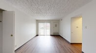 This single-floor unit accommodates four bedrooms and one bathroom within a compact, efficiently arranged floor plan. The living room serves as the central gathering space, finished with durable hardwood-style flooring and a distinctive textured ceiling. A sliding glass door allows natural light to fill the room and provides a view of the outdoors, while vertical blinds offer privacy. A built-in heating unit is positioned along the wall.

The four bedrooms are carpeted throughout and feature neutral white walls, dark baseboards, and blinds on the windows. Multiple bedrooms benefit from more than one window, allowing for natural light and outdoor views. The primary bedroom offers a modest but functional footprint. Additional bedrooms are similarly appointed, with overhead vents and standard electrical outlets.

The kitchen includes warm-toned wood cabinetry with gold hardware, white countertops, and light hardwood-style flooring. A window above the counter provides natural light and an outdoor view. The full bathroom is finished with tile flooring, a wood-tone vanity with a white countertop, a wall mirror, and a bathtub. Two hallways connect the rooms, contributing to an organized and navigable layout throughout the unit.