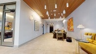 Located at 2108 Berkeley Way in Berkeley, California, this thoughtfully designed lobby space makes a strong first impression with its refined contemporary aesthetic. The entrance greets visitors through floor-to-ceiling glass double doors framed in warm wood tones, flooding the interior with natural light and offering open views to the streetscape beyond. A branded entry mat anchors the threshold, setting the tone for the curated experience within.

The expansive interior is finished with large-format, light-toned porcelain tile flooring that enhances the sense of openness. A standout architectural feature is the slatted wood ceiling, which runs the length of the space and adds warmth and texture against the crisp, light gray walls. A carefully arranged array of pendant and spot lights illuminate the ceiling, creating a layered, inviting ambiance.

The lobby is thoughtfully furnished with distinct seating vignettes. A solid wood communal table surrounded by sculptural upholstered chairs serves as a central gathering point, while a vibrant mustard-yellow banquette and teal accent chair provide a colorful lounge area near the elevator. Bold original artwork — including a large-scale abstract canvas in warm reds and oranges — adds an artistic energy to the walls. A round statement mirror and carefully placed wall sconces complete the sophisticated, gallery-like atmosphere. A connecting hallway leads deeper into the building.