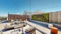 Located in the heart of downtown Wilmington, Delaware, this rooftop terrace offers an expansive outdoor retreat above the city skyline. The space is thoughtfully divided into multiple functional zones, each designed to maximize comfort and enjoyment. A covered pergola structure with slatted louvers provides adjustable shade over a well-appointed outdoor kitchen, complete with sleek countertops and cabinetry, making it ideal for outdoor dining and entertaining. Adjacent to the kitchen area, a dining table and seating provide a dedicated space for meals with a backdrop of open sky and surrounding architecture. Moving across the terrace, a dedicated lounge zone features comfortable seating arrangements, including chaise lounges and a sectional sofa surrounding a fire pit, all framed by raised planters bursting with colorful flowers and greenery. The perimeter of the terrace is lined with metal railings, allowing unobstructed panoramic views of Wilmington's cityscape, neighboring office towers, and lush treetops stretching toward the horizon. Large-format tile paving covers the entire deck surface, giving the space a clean, modern aesthetic. Access is provided via an elevator and stairwell entry point integrated into the building's brick facade. String lighting overhead adds a welcoming ambiance, making this rooftop an exceptional urban outdoor destination.