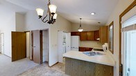 This single-floor residence offers a well-designed layout centered around a functional and inviting kitchen. The kitchen is highlighted by warm oak cabinetry that lines the walls, providing ample storage alongside a suite of white appliances. A peninsula extends into the space, complete with a stainless steel sink and a light-toned countertop, offering both additional workspace and a natural division between the kitchen and adjoining areas. Overhead, vaulted ceilings lend an open, airy quality to the space, enhanced by a classic wrought-iron chandelier with frosted glass shades and a matching pendant light positioned above the peninsula. Recessed lighting further brightens the area. The flooring features neutral tile that complements the overall warm, understated color palette throughout. A dedicated pantry or utility area is visible nearby, featuring built-in shelving and space for a washer and dryer, adding to the home's practicality. A door leads to additional storage, including what appears to be a closet with louvered doors. The overall ambiance is warm, comfortable, and functional, making this single-floor home well-suited for everyday living.