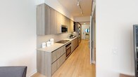 Located at 1201 Half St SW in Washington DC's vibrant Southwest Waterfront neighborhood, this thoughtfully designed single-floor residence offers a refined blend of contemporary style and functional living. Light oak hardwood flooring flows throughout, tying each space together seamlessly.

The open kitchen features floor-to-ceiling flat-front cabinetry in a warm wood-tone finish, complemented by stainless steel appliances including a built-in microwave, oven, and refrigerator. Light quartz countertops and track lighting overhead complete the sleek, modern aesthetic.

The living and dining area is anchored by expansive floor-to-ceiling windows that flood the space with natural light and offer dynamic city views. A muted blue accent wall provides a striking backdrop for a curated gallery arrangement, adding personality and visual depth to the room.

The bedroom is comfortably appointed with wall-mounted sconce lighting flanking a tufted upholstered headboard, alongside warm wood furniture that creates an inviting, restful atmosphere. The bathroom continues the modern design language with large-format tile, a floating vanity, and a full soaking tub with overhead rain-style shower. Clean lines and neutral tones throughout lend the entire space a cohesive, polished character.